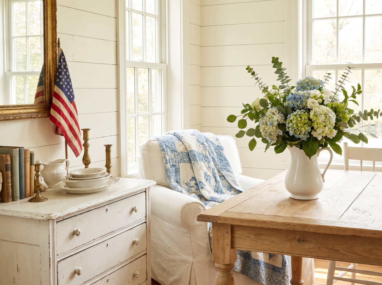 A cozy light cottage corner with a blue and white quilt, vintage American flag, light wood antiques and hydrangeas