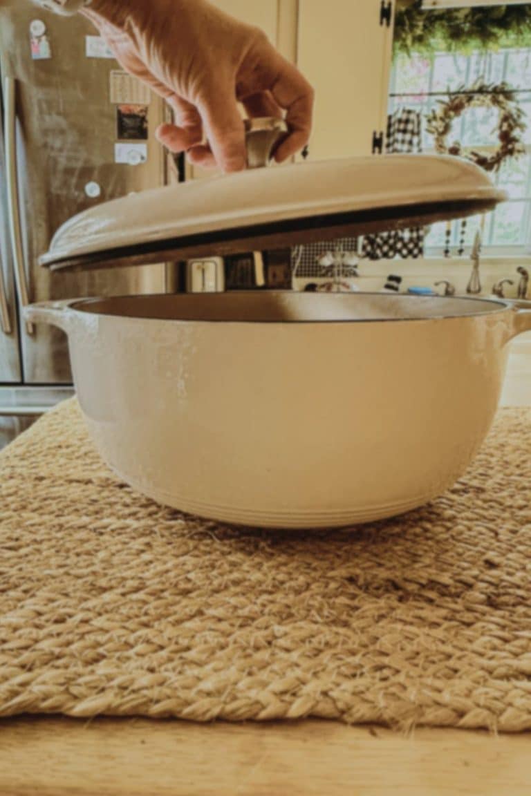Hand lifting the lid of a cream enameled cast iron Dutch oven on a woven jute mat in a farmhouse kitchen