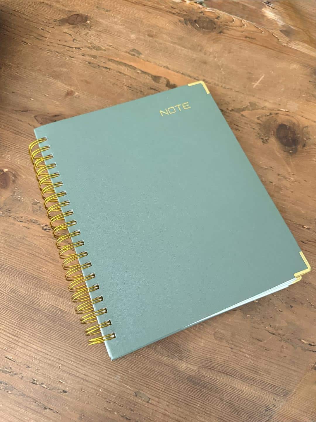 Olive green spiral notebook with gold wire binding and brass corners on weathered barnwood in warm afternoon light
